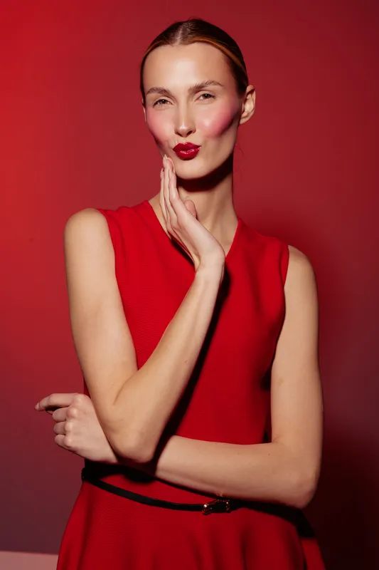 Young woman in red dress poses with hand on face and red lipstick on red background. Concept of fashion, beauty, makeup, and confident female model portrait.