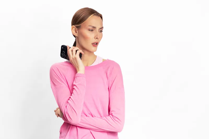 Young woman in pink sweater holds smartphone near ear, looking concerned and thoughtful on white background. Concept of communication, decision making, and uncertainty.