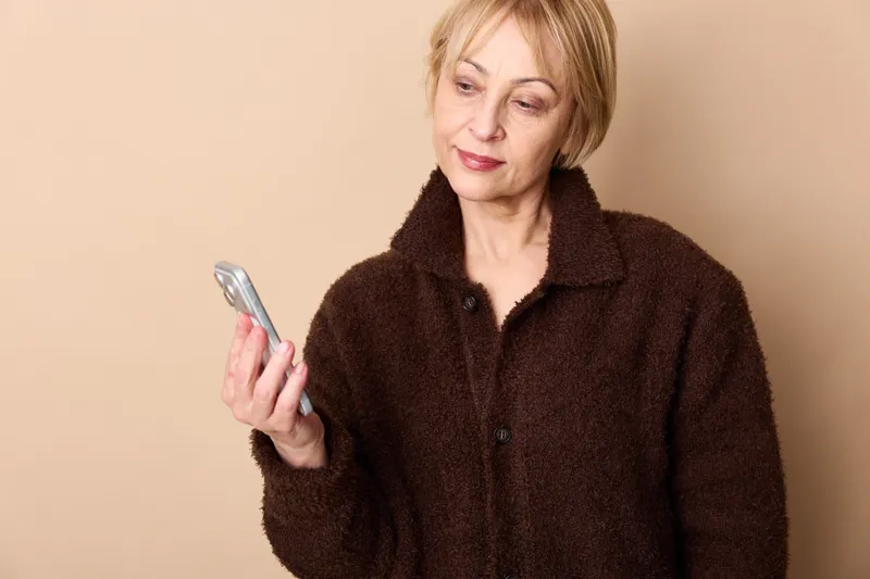 Smiling senior woman, blond hair, holding smartphone, looking at screen, indoor portrait on beige background, relaxed mood, technology communication
