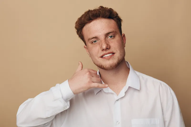 Young man in white shirt makes call me gesture with hand near face, smiling gently on beige background. Concept for communication, phone call, casual lifestyle and friendly gesture.