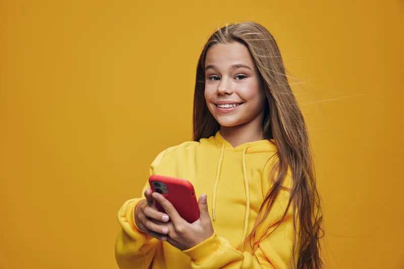 Girl with long hair smiling and holding smartphone in red case wearing yellow hoodie on bright orange background for social media or communication.