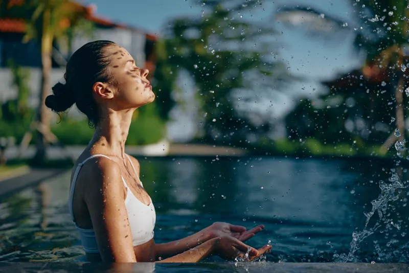 Cheerful woman outdoors in a pool, eyes closed, sunlit moment, water spray around, relaxed posture, vibrant greenery background, refreshing summer mood and natural light