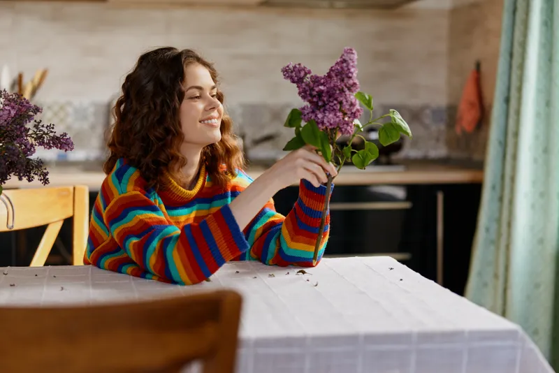 Smiling woman with curly hair in a striped sweater examines a lilac bouquet at a bright kitchen table, capturing a casual home moment of comfort, self care, and everyday joy.