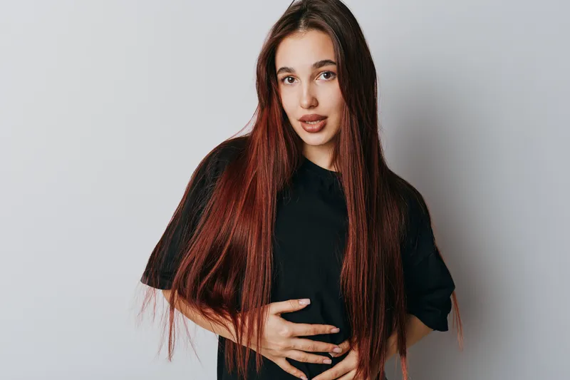Young woman with long straight hair and natural makeup wearing black t shirt standing with hands on stomach against white background. Portrait of female model with confident expression.