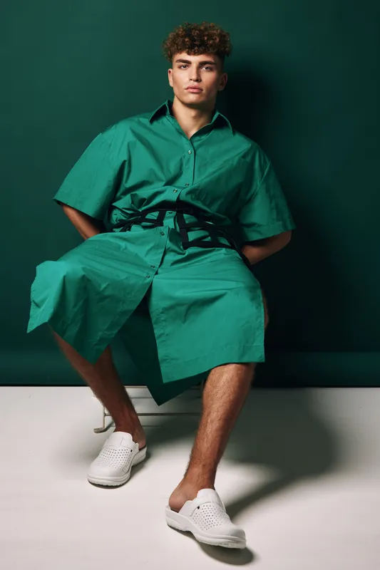 Young man with curly hair wearing green oversized shirt dress and white slip on shoes sitting on chair against dark green background in studio.