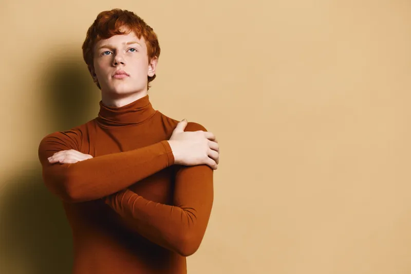 Confident redhead young man in brown turtleneck sweater standing with crossed arms against neutral background with thoughtful expression and casual style.