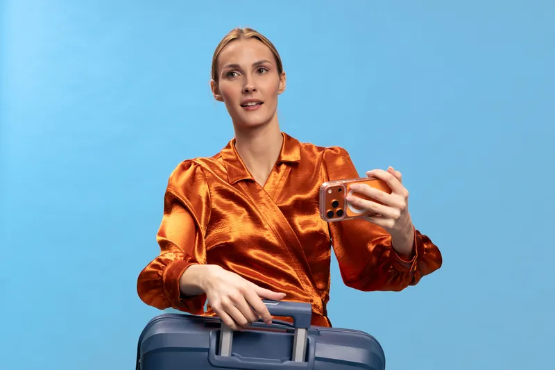 Woman in orange satin blouse holds phone and suitcase on blue background. Travel concept with female traveler preparing for trip and casual expression.