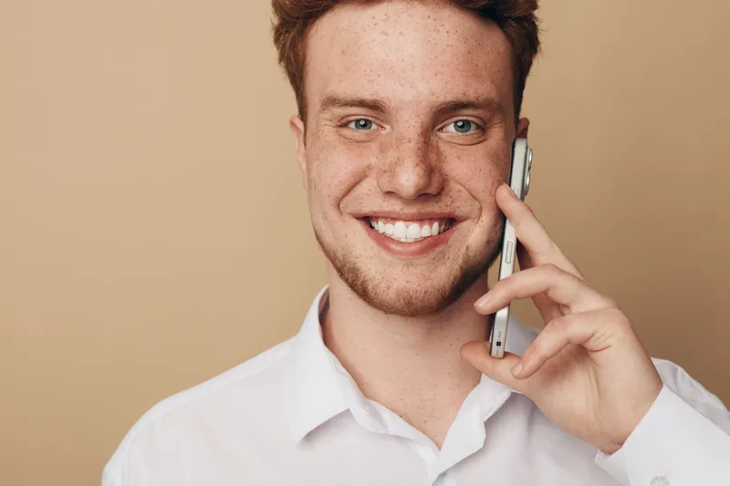 Smiling young man with red hair and freckles talks on smartphone. Casual white shirt and beige background create friendly and positive communication concept in modern lifestyle.