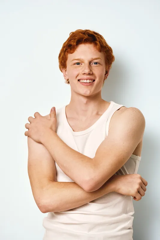 Young man with red hair and freckles smiling wearing sleeveless white shirt standing against white background in studio. Portrait with crossed arms and natural light.