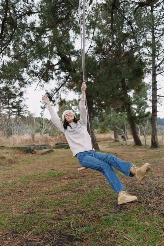 Woman on a swing in a forest clearing enjoying outdoors, wearing blue jeans and a sweater, laughter and freedom among tall trees and fresh air.