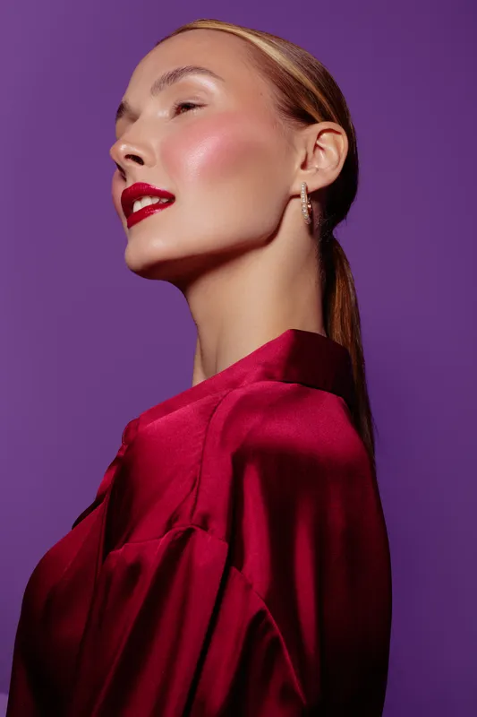 Profile of a confident woman with red lipstick and shiny skin wearing a red satin blouse against purple background. Concept of beauty, fashion, elegance, and makeup.