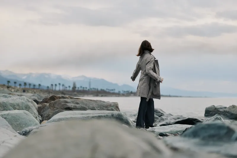 A woman in a long coat stands on rocky shore looking at the sea under cloudy sky. Mountains and palm trees are visible in the background. Concept of solitude and nature.