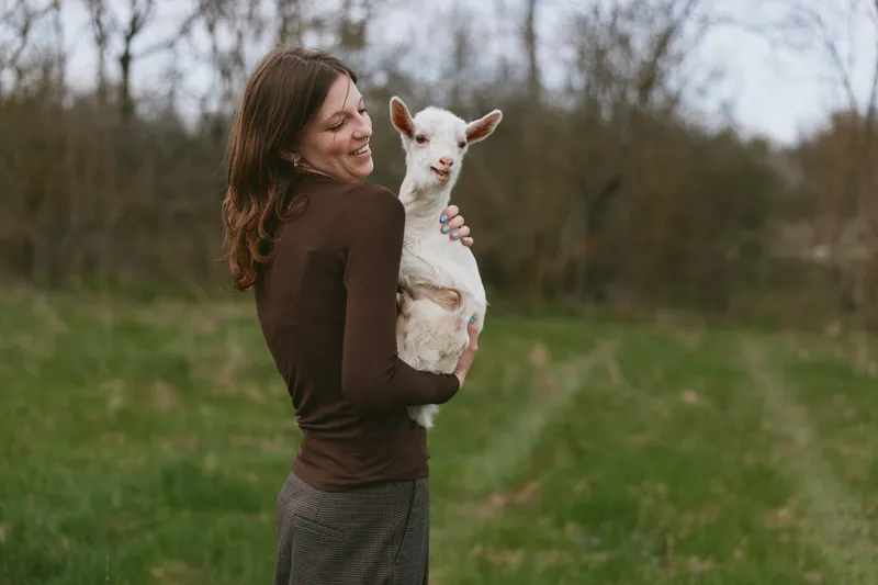 Young woman holds a white baby goat in a green field during autumn while smiling and enjoying a peaceful moment outdoors.