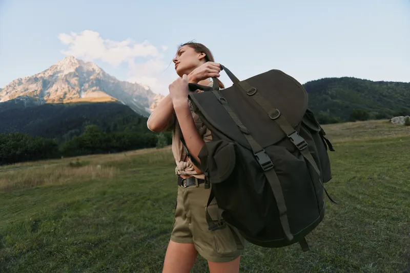A woman with a large backpack stands in a meadow with mountains in the background, preparing for a hike. An outdoorsy lifestyle moment emphasizing adventure and travel.