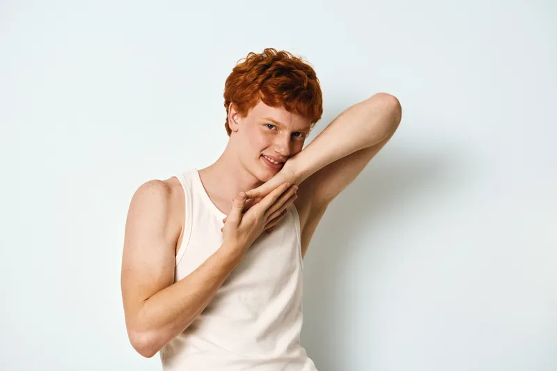 Young man with red hair in white sleeveless shirt posing with hands near face against plain white background in studio for portrait photography.