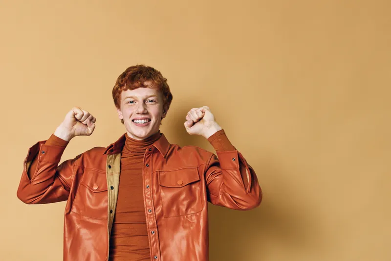 Young man with red hair smiling and raising fists in celebration wearing orange jacket and turtleneck standing against beige background for success and happiness.