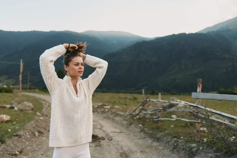 Young woman in a white knit sweater stands on a mountain road, adjusting her hair with a calm, contemplative expression.