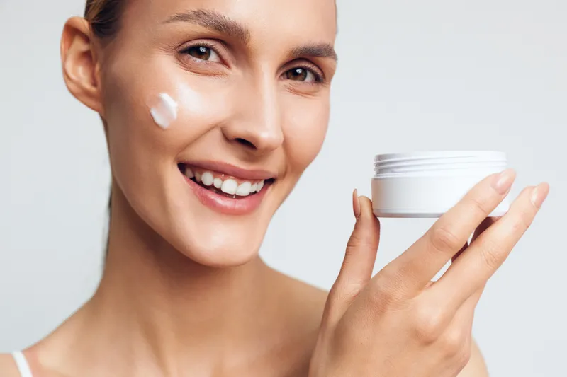 Smiling woman applying white face cream on cheek holding jar with skincare product. Close-up on light background. Concept of beauty, wellness, and daily self-care routine.