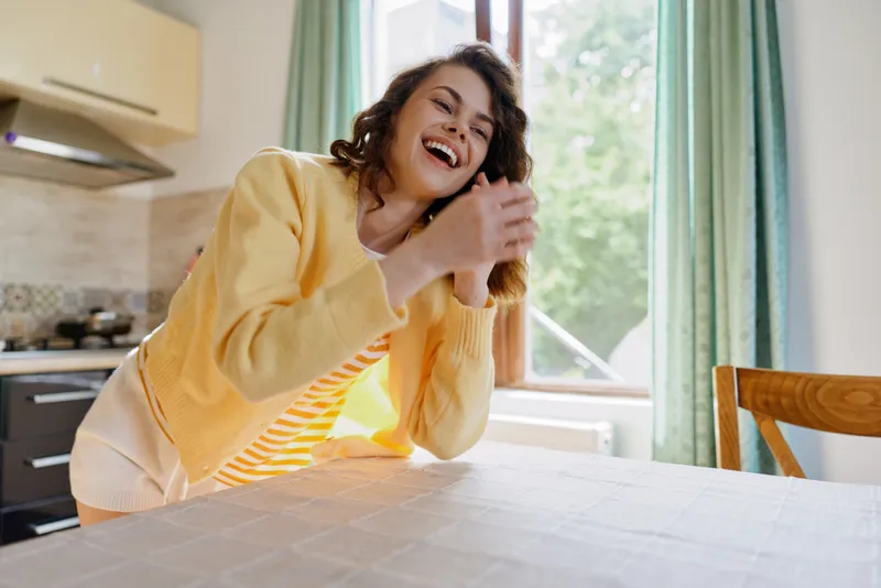 Smiling woman in a yellow cardigan leaning on a kitchen table with bright morning light, a warm home scene conveying casual lifestyle, comfort and wellness