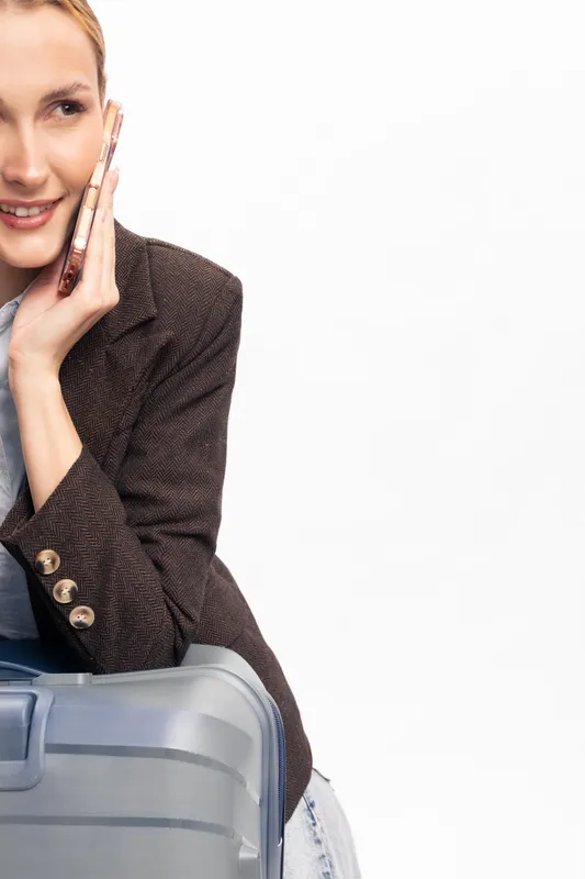 Smiling young woman talks on smartphone while sitting on suitcase. Casual female traveler wears brown jacket and light jeans. Concept of travel, communication, and leisure time.