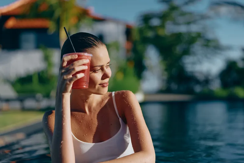 Relaxed woman enjoys a refreshing juice by the pool on a sunny day, wearing a white tank top, leaning on the edge, savoring the moment, demonstrating casual lifestyle and outdoor leisure by vibrant