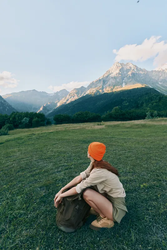 Woman sits in a wide grassy field with a backpack and orange beanie, enjoying a peaceful outdoor lifestyle amid distant mountains and expansive nature scenery.