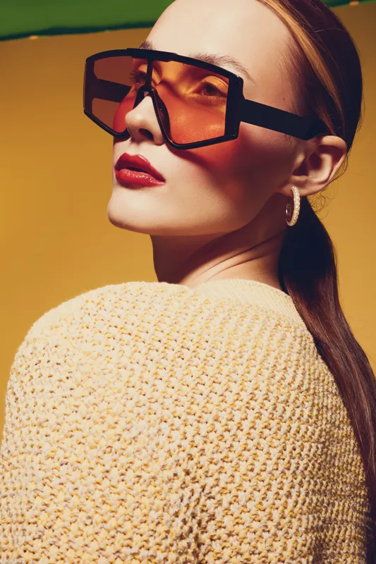 Stylish woman with large orange sunglasses and red lipstick poses against yellow background. Fashion portrait with knitted sweater and hoop earrings in warm studio lighting.