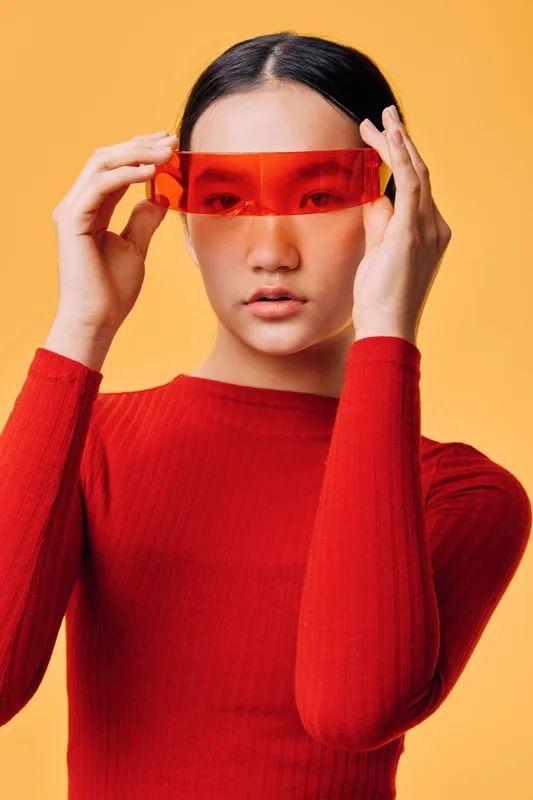 Woman with black hair wearing red long sleeve shirt holding translucent red visor against orange background. Portrait with futuristic fashion style and minimal makeup on face.