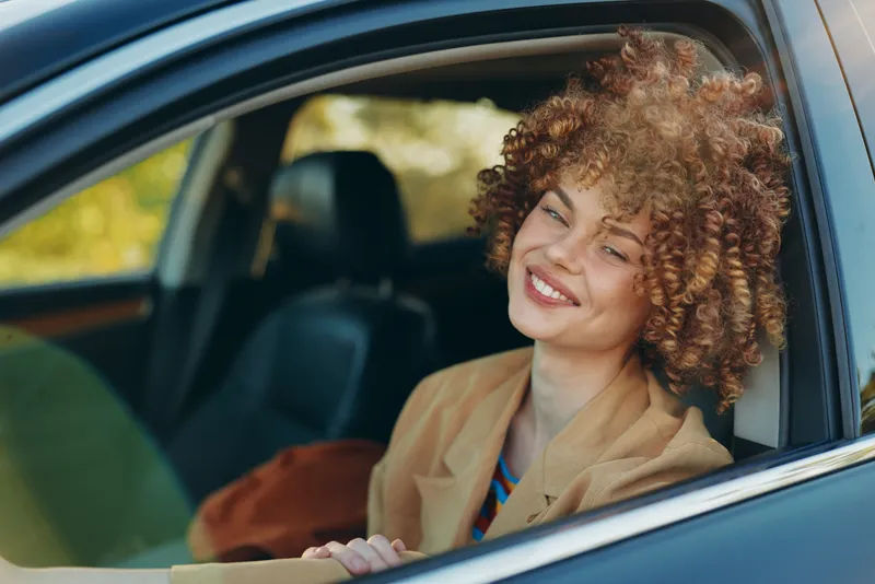 Woman in a car smiling, casual lifestyle scene with retro film color grading, warm natural light, relaxed pose, everyday commute mood, bright happiness captured