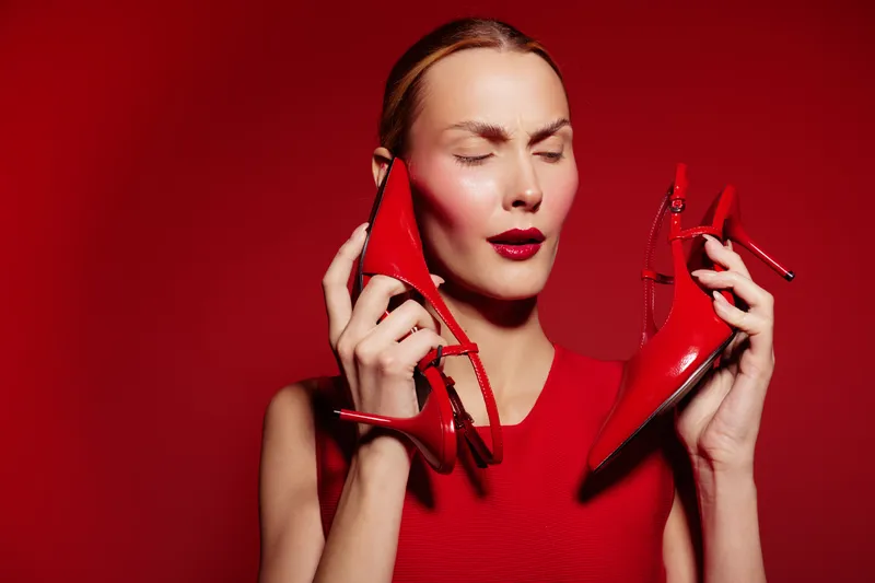 Woman in red dress holds red high heels near face with closed eyes on red background. Concept of fashion, style, elegance, and bold color contrast in studio.
