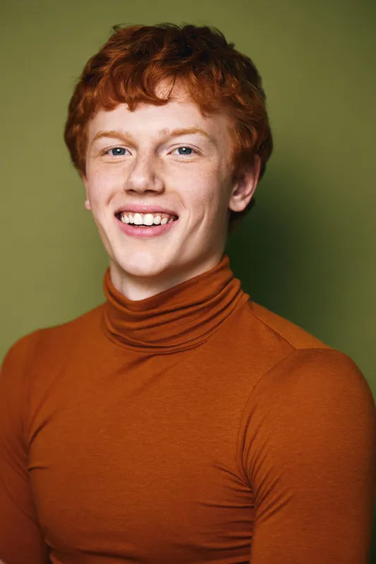 Portrait of smiling young man with red hair wearing orange turtleneck against green background. Happy male model posing with natural expression and bright eyes in studio photo.