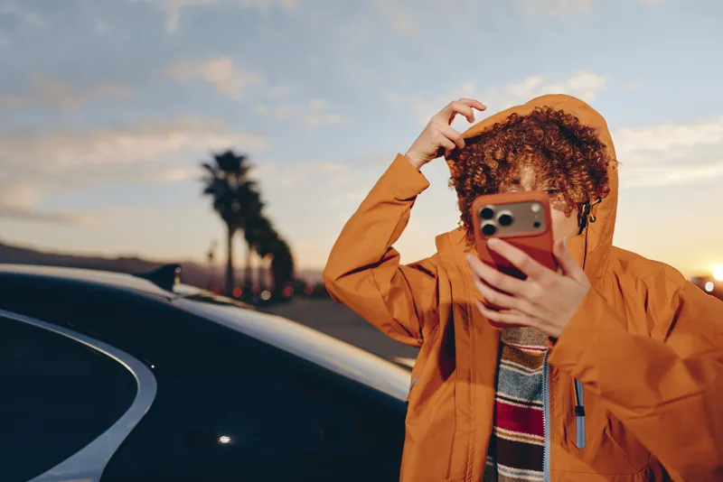 Woman lifestyle portrait, holding a smartphone for a selfie, curly hair under an orange jacket, standing by a car at sunset, outdoor travel mood, warm golden light, soft sky background