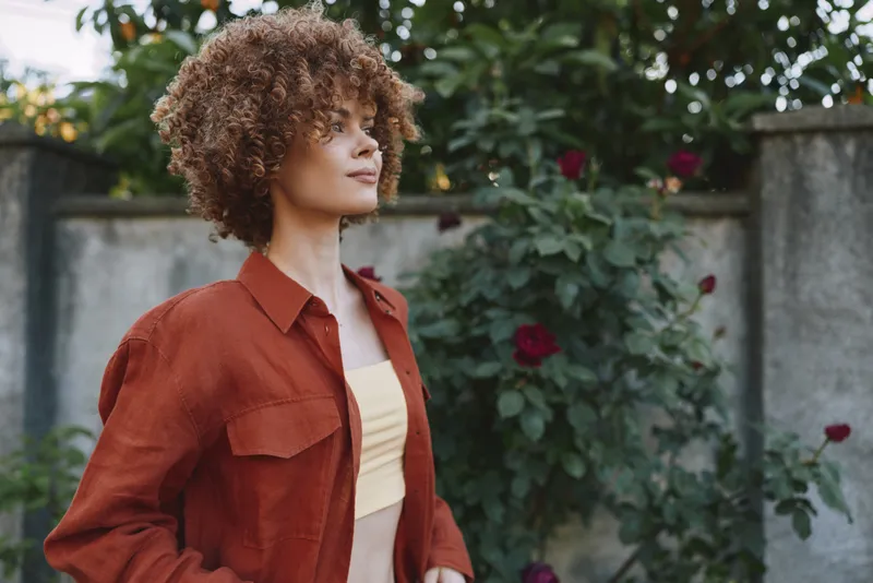 Woman with curly hair in rust shirt stands in a garden, casual lifestyle portrait, natural daylight, soft smile, roses in background, outdoor serene mood