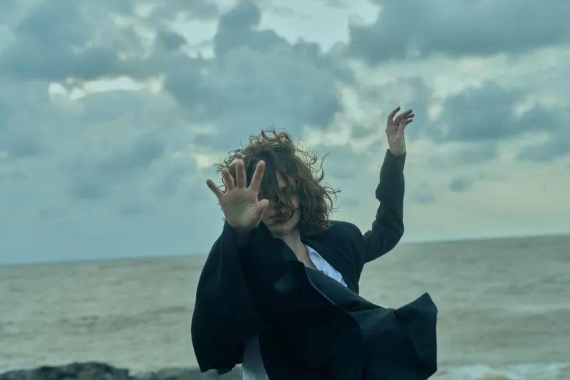 Windy beach scene with a figure in motion, a coat flaring, hands raised, sea and clouds behind, conveying energy, drama, and escape from stillness in a dynamic outdoor moment