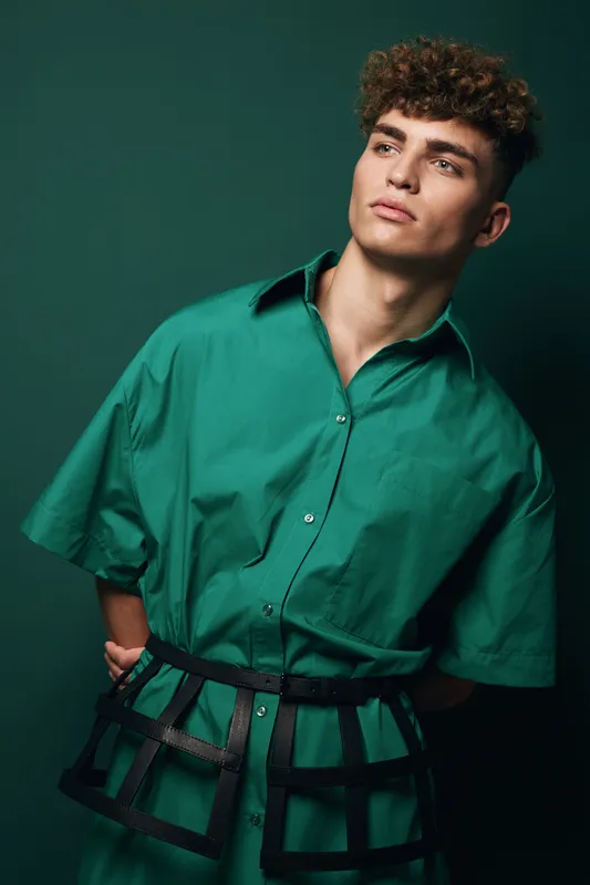 Young man with curly hair wearing oversized green shirt and black harness posing against dark green background in studio for fashion portrait and style concept.