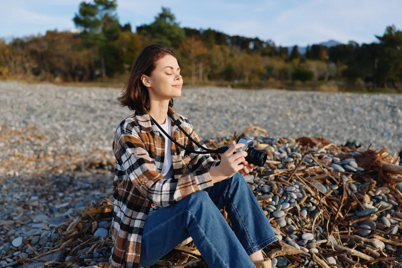 Woman with camera sits on a pebble beach, basking in sunlight, enjoying outdoor photography and nature, capturing landscape textures with calm focus amid tranquil shoreline scenery.