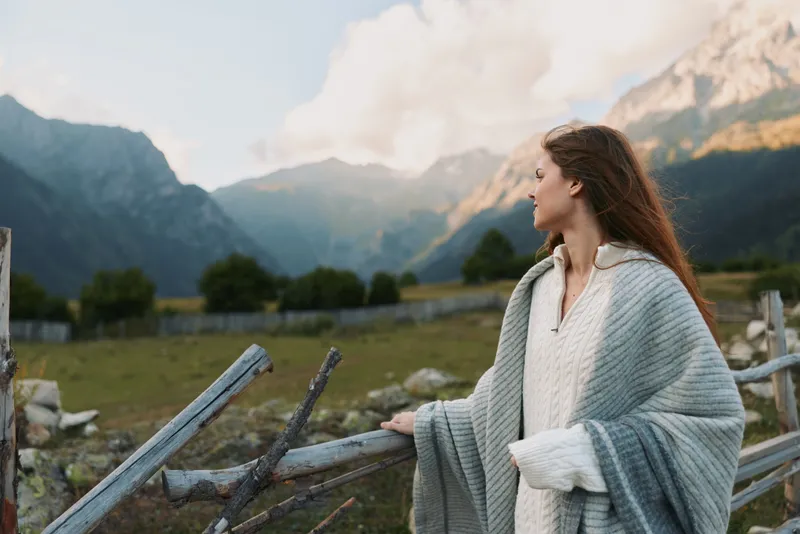 Young woman wrapped in cozy blanket stands by wooden fence looks at mountain landscape enjoys peaceful nature outdoors in autumn day lifestyle concept.