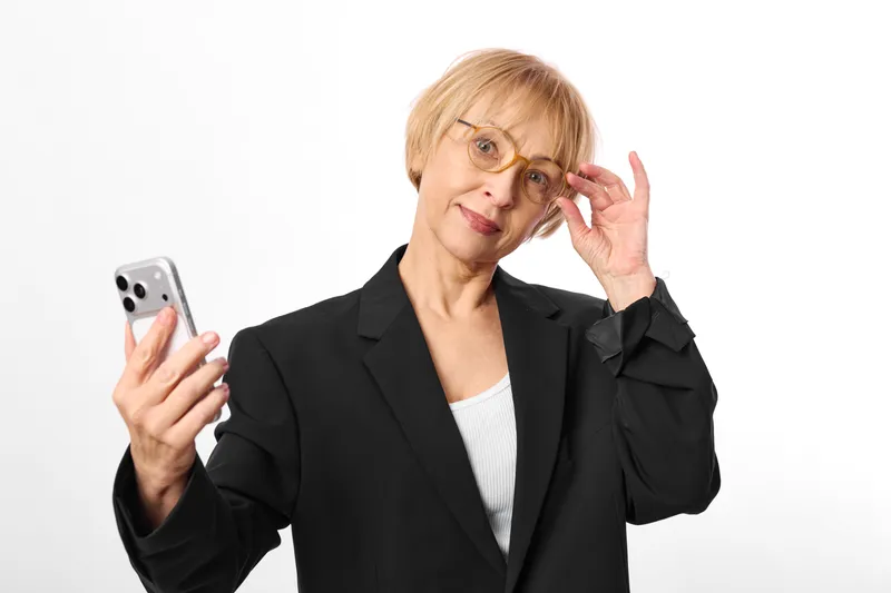 Senior woman with smartphone in studio portrait, thoughtful smile, raising hand near face, communication gesture, modern device, black blazer, clean white background, soft light