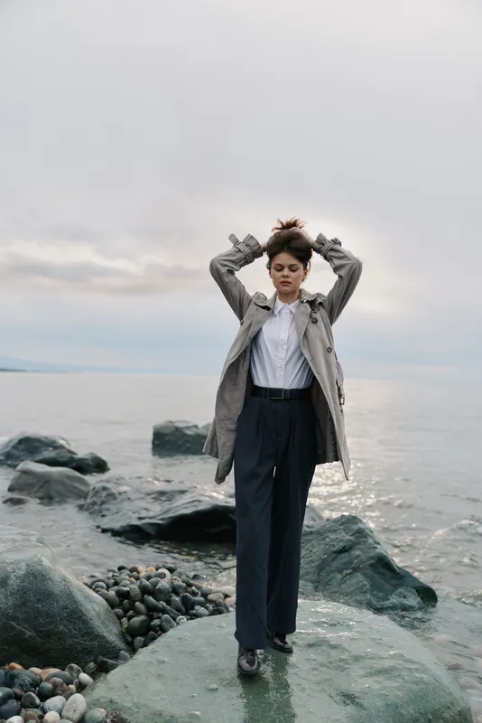 Young woman in stylish coat and white shirt stands on rocky shore with calm sea and cloudy sky background. Concept of fashion, nature, and peaceful outdoor lifestyle.