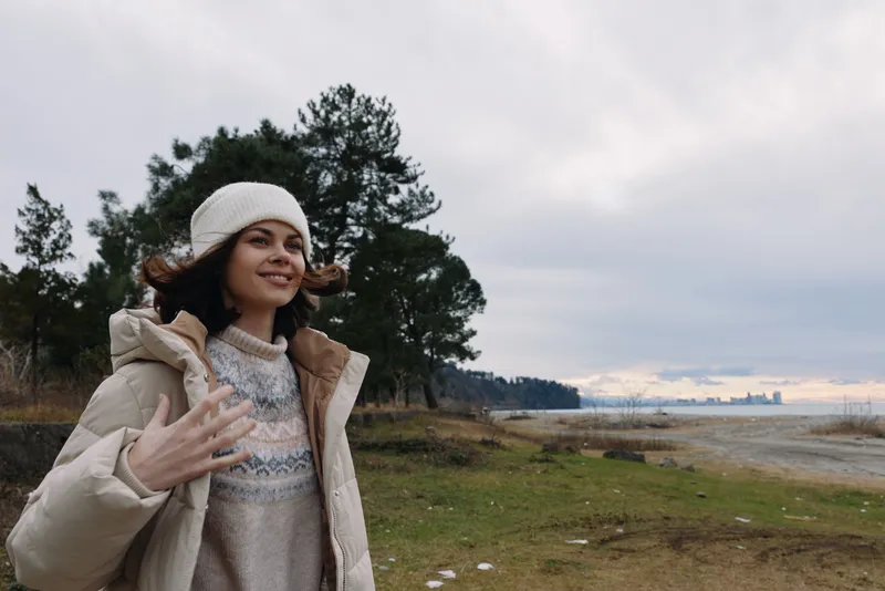 Woman outdoors in a warm coat on a windy day by a grassy coast with trees and a distant water view showcasing nature and calm mood