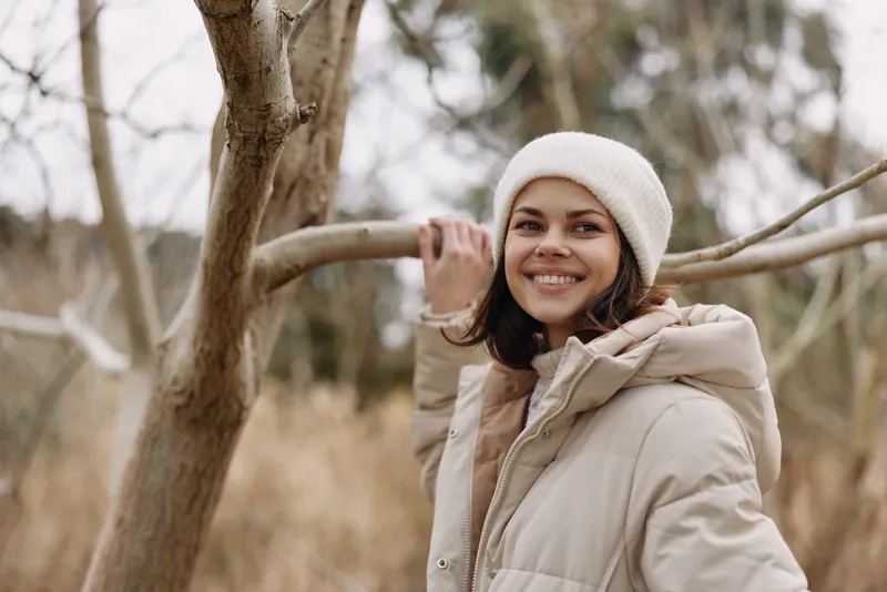 Woman in a beige coat and white beanie smiles while standing among bare trees in a park, capturing a warm, candid moment of outdoor style and seasonal calm in soft light.