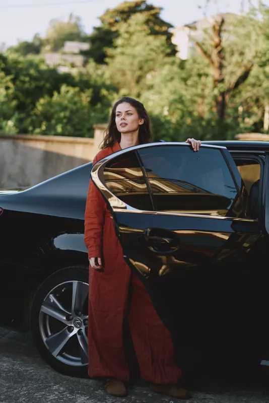 Woman lifestyle outdoors by a sleek car with open door, wearing a red cardigan, relaxed pose, sunlight and breeze, natural colors with greenery in the background