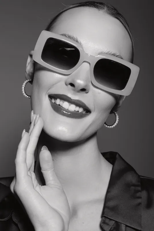 Smiling Caucasian woman with fair skin wearing large sunglasses and hoop earrings touches her face. Studio portrait with fashionable accessories and positive mood.