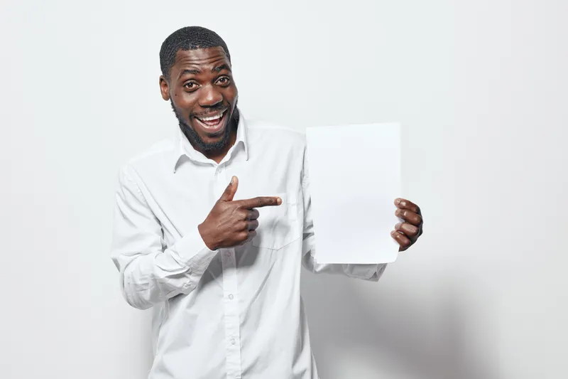 African American man with dark skin wearing white shirt smiling and pointing at blank white paper for presentation or advertisement on white background.