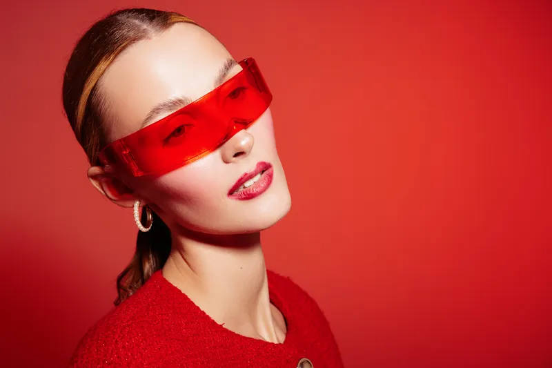 Young woman with red sunglasses and red sweater poses against red background. Fashion portrait with glossy lips and hoop earrings. Concept of modern style and bold color contrast.