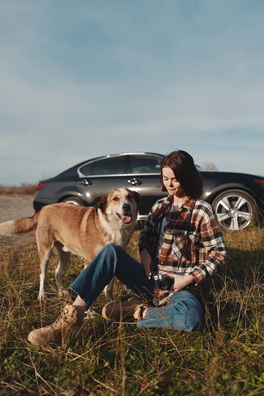 Dog, woman, outdoors, car, grass, pet, companionship, leisure, nature, field, relaxed pose on sunny day with dog beside woman in plaid shirt and jeans in open countryside