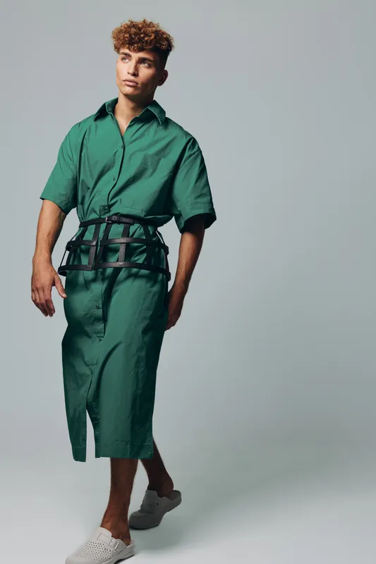Young man with curly hair wearing green shirt dress and black belt walking barefoot on light background. Fashion model posing in casual outfit with relaxed expression and modern style.