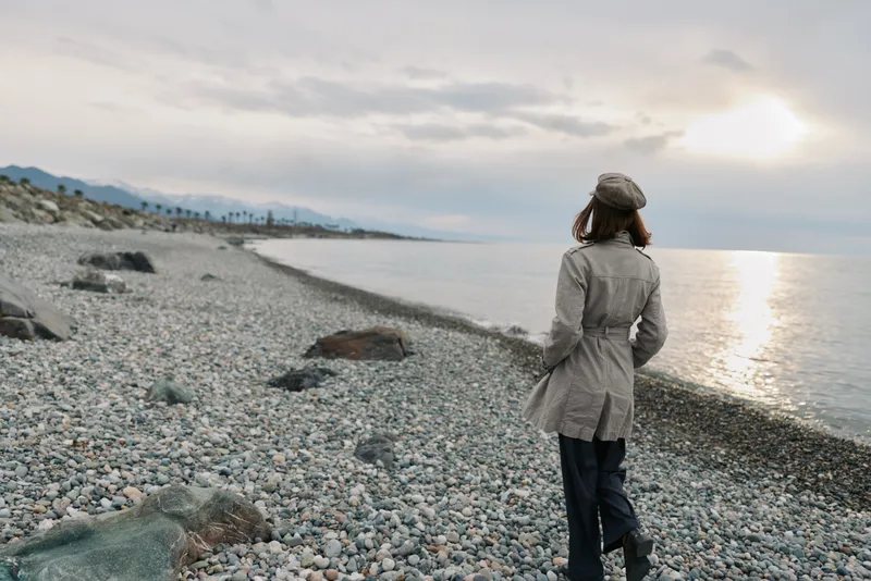 A woman in a coat and beret walks alone on a rocky beach during sunset. The calm sea and cloudy sky create a peaceful and contemplative atmosphere by the shore.