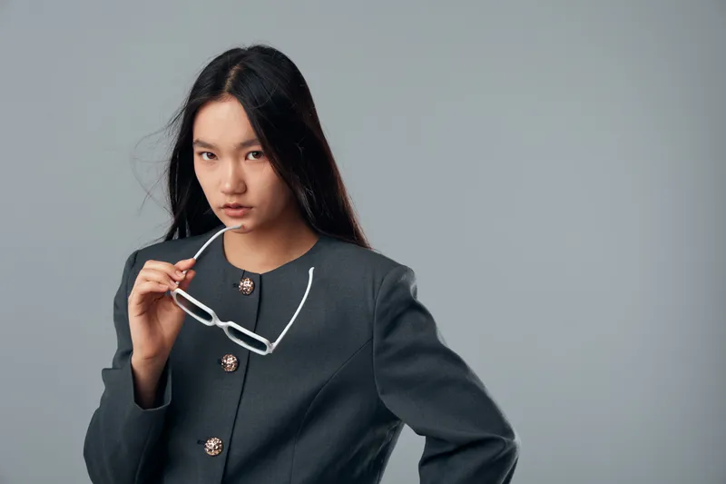 Young woman with long black hair wearing dark gray blazer holding white glasses with confident expression on gray background, studio portrait with minimalistic style and fashion details.