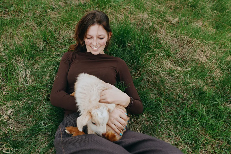 Young woman with long brown hair lies on green grass holding a small white baby goat while smiling and enjoying a peaceful outdoor moment in casual autumn clothing.